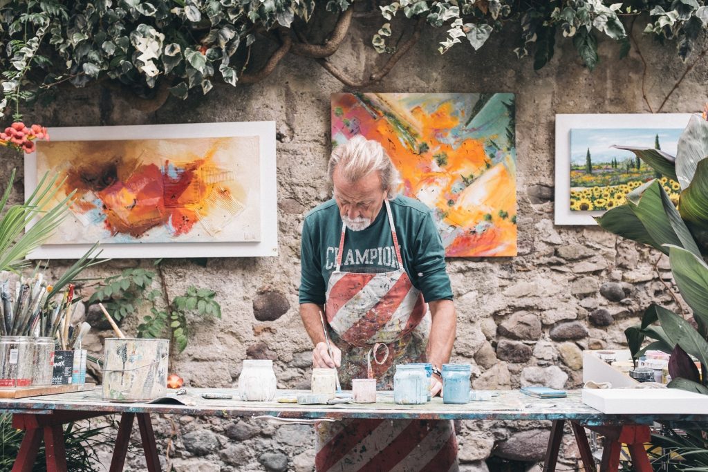 a senior artist painting outside with paintings hung up